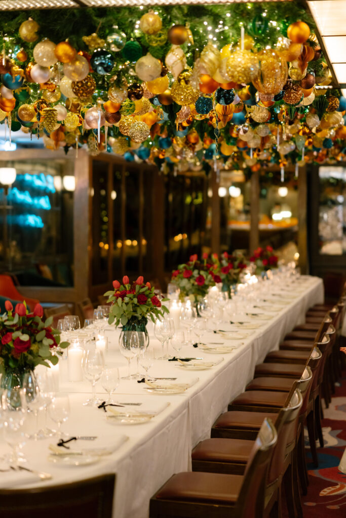 A long, elegant dining table is set with white linens, floral centerpieces, and glassware. Above, colorful ornaments and greenery hang from the ceiling, creating a festive atmosphere in a warmly lit room.