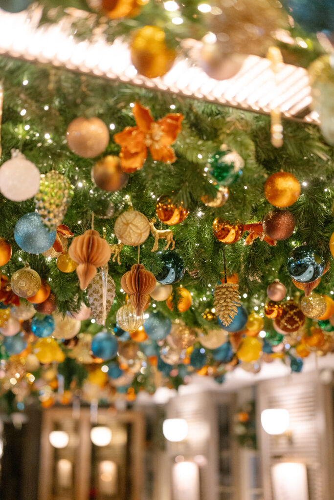 A festive ceiling decorated with green garland and assorted colorful ornaments, including gold, blue, and orange balls, paper decorations, and pinecones, with warm lights glowing in the background.