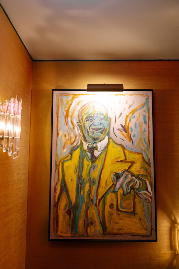 A colorful, expressive painting of a man in a yellow suit hangs on a warmly lit, tan wall. The figure points outward, and the style is abstract and bold. A light fixture above illuminates the artwork.