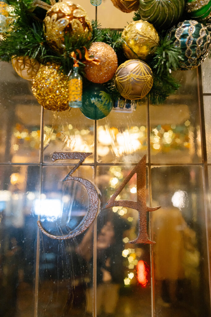 A glass door with the number 34 displayed in metallic letters, decorated with a festive wreath of green pine, gold, green, and blue ornaments, and twinkling lights reflected in the background.