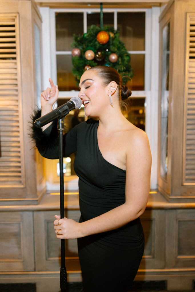 A woman in a one-shoulder black dress sings passionately into a microphone indoors, standing in front of a window decorated with a festive wreath.