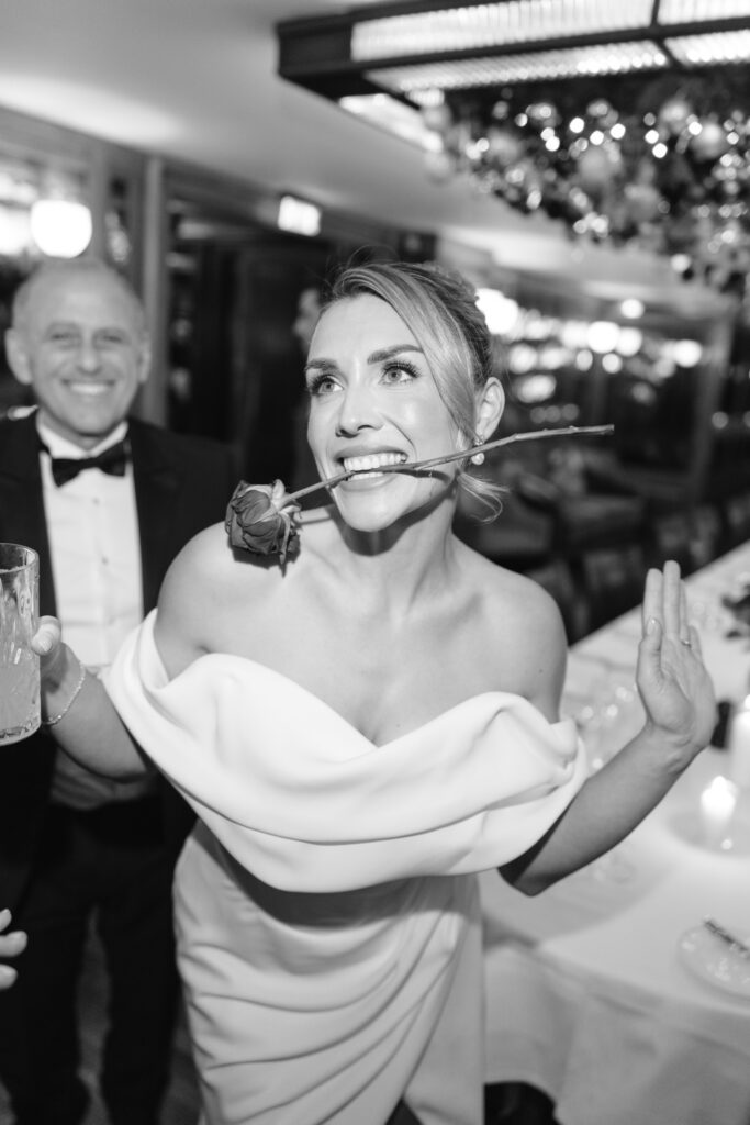 A woman in an off-the-shoulder dress smiles playfully with a rose in her mouth, holding a drink and raising one hand. A man in a tuxedo stands behind her, smiling. The scene is lively and festive.