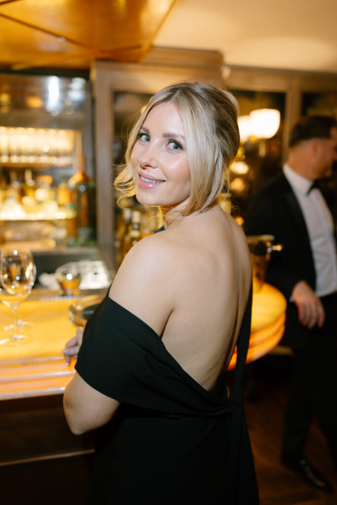 A woman in a black off-the-shoulder dress stands at a bar, smiling over her shoulder at the camera. The background shows shelves with bottles and glasses, and a man in a suit is blurred in the background.