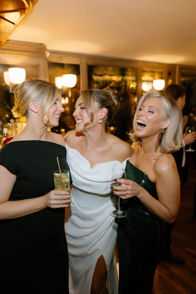 Three women dressed elegantly in evening gowns stand close together, smiling and laughing while holding drinks at a lively indoor event. Warm lighting and a festive atmosphere are visible in the background.