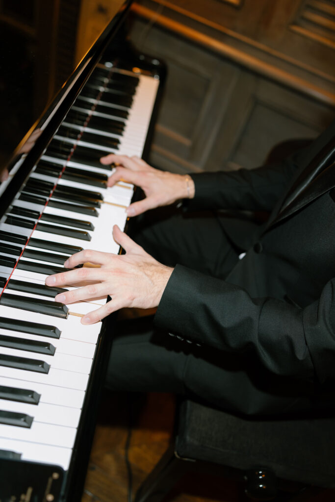 A person in a black suit plays a piano, with both hands on the keys. The wooden floor and paneling are visible in the background.