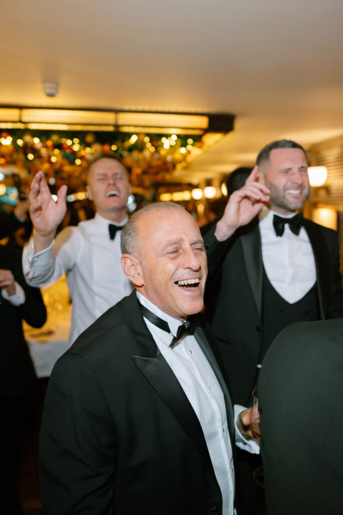 Three men in tuxedos laugh and sing together at a lively indoor event, with festive lights and decorations in the background. One man holds a drink while the others gesture cheerfully.