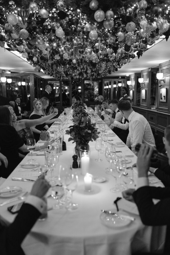 A large group of people in formal attire sit around a long, elegantly set dinner table, raising glasses for a toast under a ceiling decorated with numerous hanging ornaments and lights.