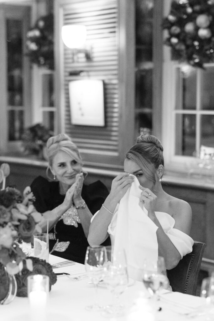 A woman in a white dress wipes her eyes with a napkin, appearing emotional, while another woman beside her claps supportively at a decorated, candlelit table in a warmly lit room.