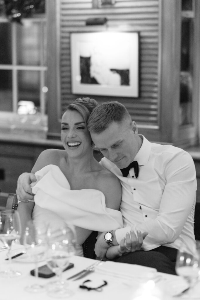A joyful bride and groom sit closely together at a table, laughing and holding hands, during a wedding reception. The image is in black and white, with a warm, intimate atmosphere.