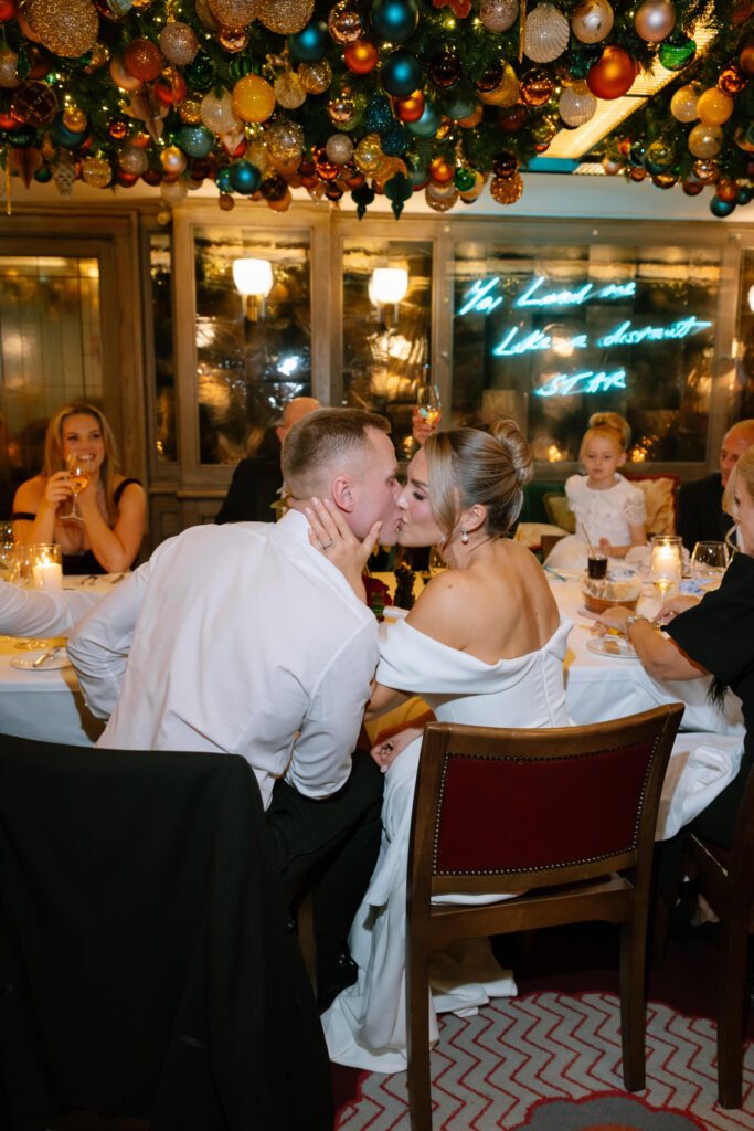 A couple in formal attire shares a kiss at a festive dinner table, surrounded by guests and colorful holiday decorations hanging from the ceiling. Warm lights and a cozy atmosphere fill the room.