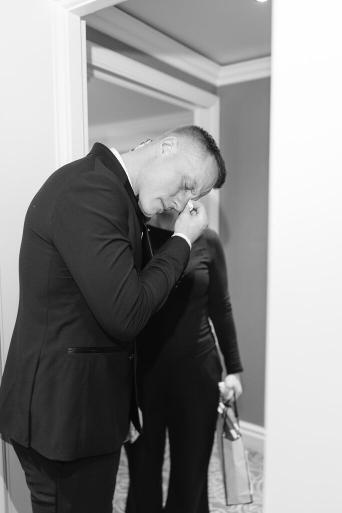 A man in a suit wipes his eye emotionally while standing in a doorway; a person in the background holds a gift bag. The scene appears to be candid and heartfelt.