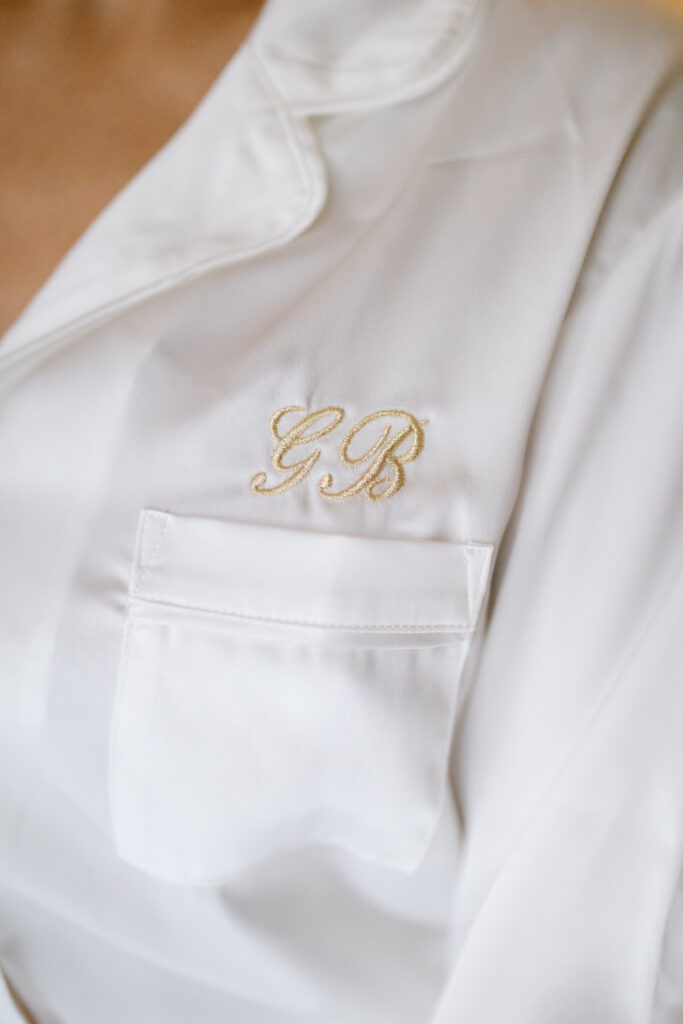 Close-up of a white satin pajama top with embroidered initials GB in gold above the chest pocket.