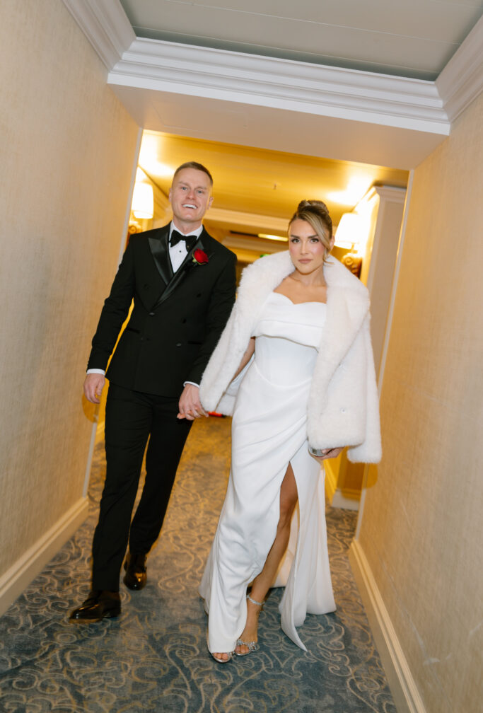 A couple walks down a hallway dressed in formal attire; the man wears a black tuxedo with a red rose, and the woman wears a white gown with a fur shawl, holding hands and looking confidently ahead.