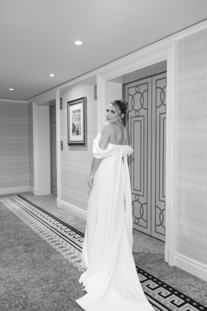A woman in an elegant, off-the-shoulder gown stands in a hallway, looking back over her shoulder. The hallway features patterned carpet, framed art, and double doors. The image is in black and white.