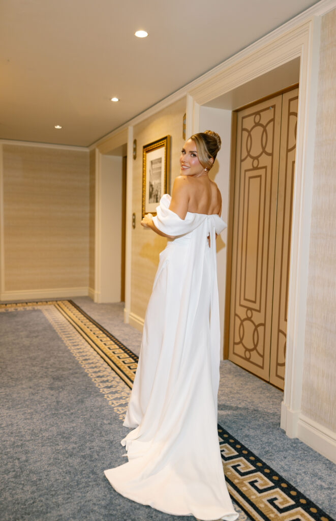 A woman in an elegant off-the-shoulder white gown stands in a carpeted hallway, looking back over her shoulder and smiling. The hallway features artwork, patterned carpet, and decorative doors.