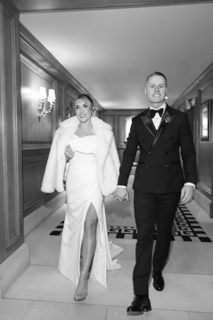 A bride in a white dress and fur jacket walks hand-in-hand with a groom in a black tuxedo down a hallway, both smiling confidently.