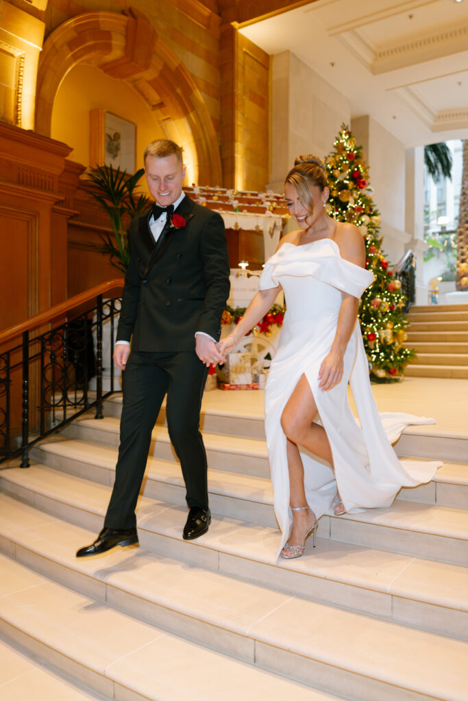 A couple dressed in formal attire, the man in a tuxedo and the woman in an off-the-shoulder white gown, walk down indoor stairs, holding hands. A decorated Christmas tree is in the background.