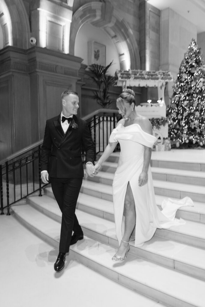 A couple in formal attire walks down elegant staircase; the man wears a tuxedo, and the woman wears a white gown with a high slit. A decorated Christmas tree and festive decorations are visible in the background. The photo is black and white.