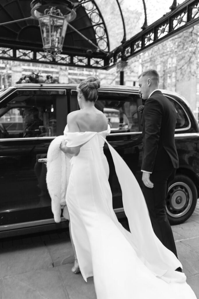A bride in an off-shoulder white gown and a groom in a black suit walk towards a classic black car under an ornate canopy. The scene is elegant and candid, likely taken on their wedding day.