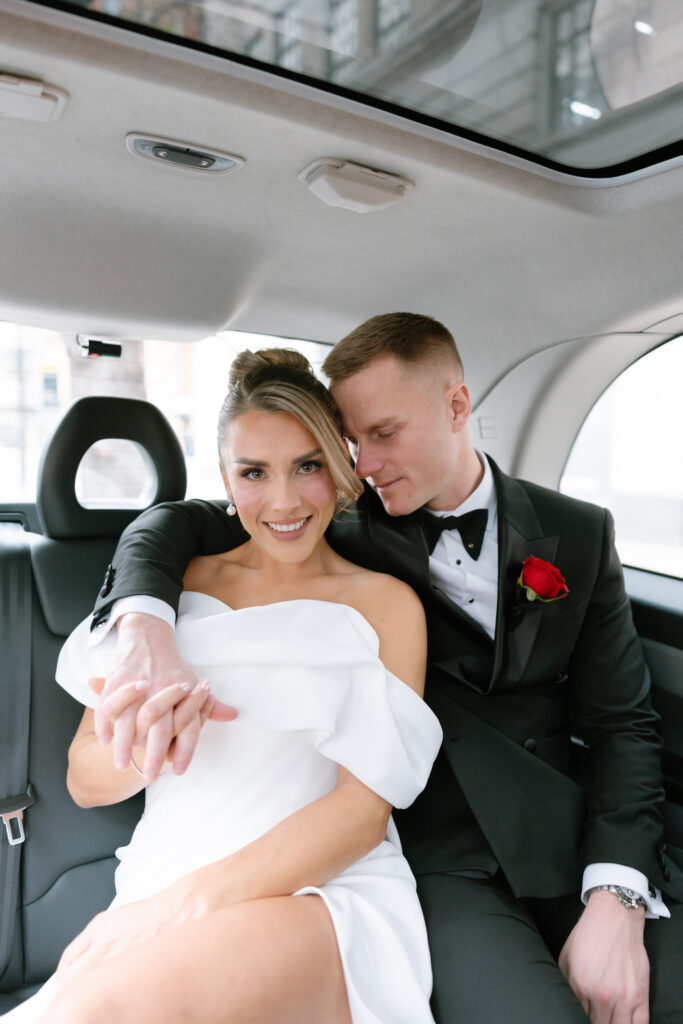 A bride in an off-the-shoulder white dress smiles while holding hands with a groom in a black tuxedo with a red rose, sitting close together in the back seat of a car.