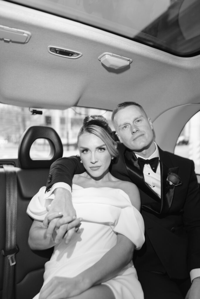 A bride in an off-shoulder white dress and a groom in a tuxedo sit closely together in the backseat of a car, holding hands and looking confidently at the camera.