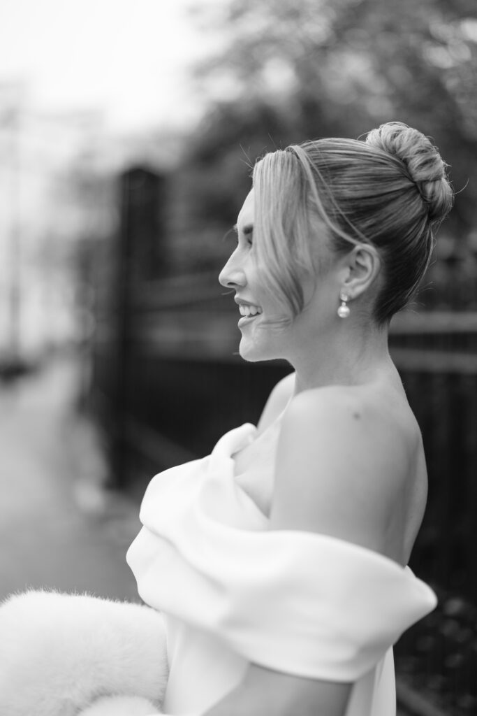 A black-and-white photo of a smiling woman in an off-the-shoulder dress, with her hair in a bun and pearl earrings, standing outdoors near a fence.