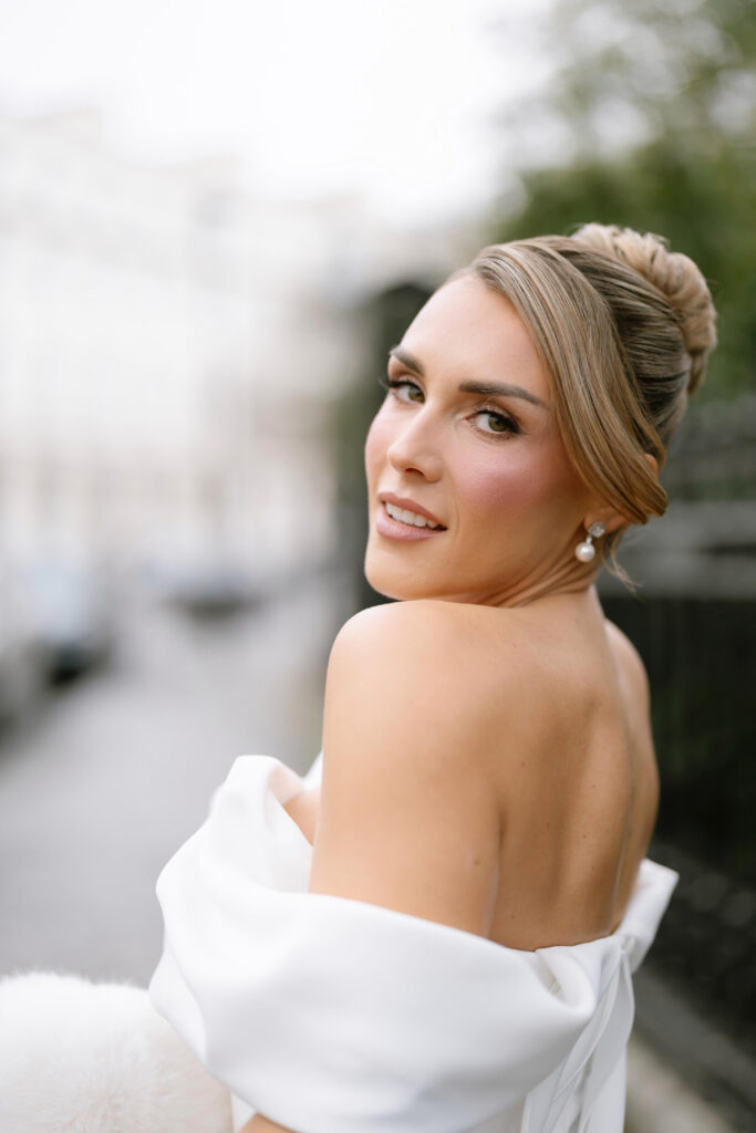 A woman with styled blonde hair and pearl earrings wears an off-the-shoulder white dress, looking back over her shoulder and smiling softly outdoors. The background is blurred with hints of buildings and greenery.