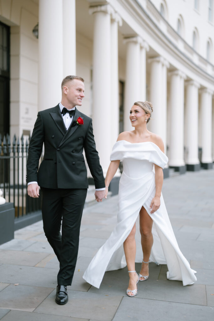 A couple in formal attire walks hand in hand outdoors. The man wears a black tuxedo with a red rose boutonniere, and the woman wears an off-the-shoulder white gown with a high slit. Both are smiling and looking at each other.