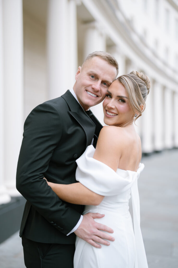 A smiling couple in formal attire embraces outdoors. The woman wears an off-shoulder white dress, and the man wears a black suit. White columns and a curved building appear in the background.