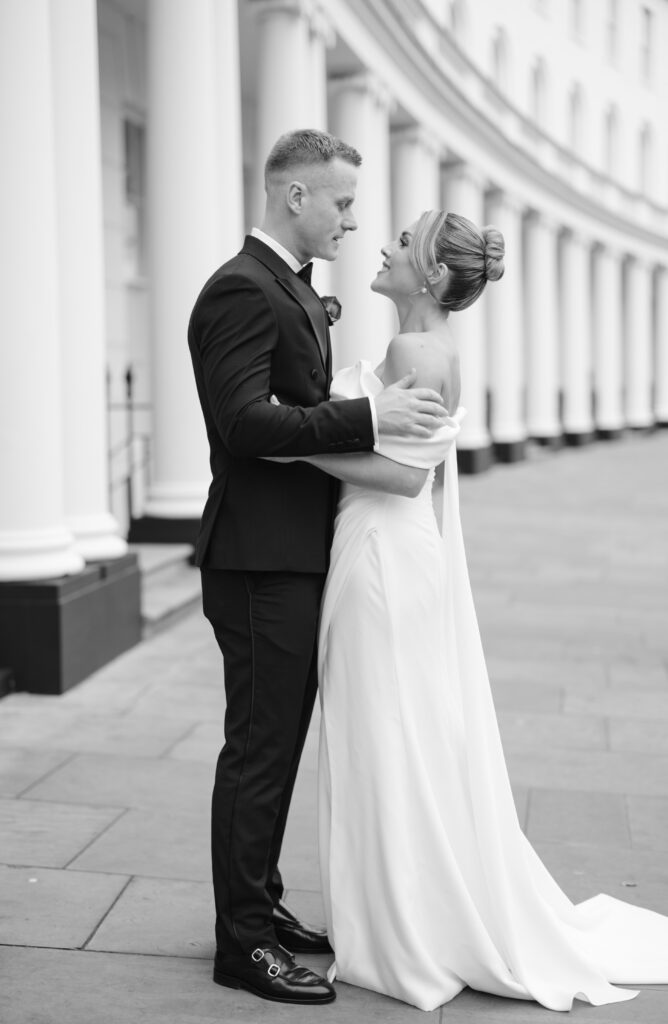 A bride and groom stand facing each other, embracing and smiling, in front of a row of white columns on a city sidewalk. The bride wears a long, elegant gown and the groom wears a dark suit. The image is in black and white.