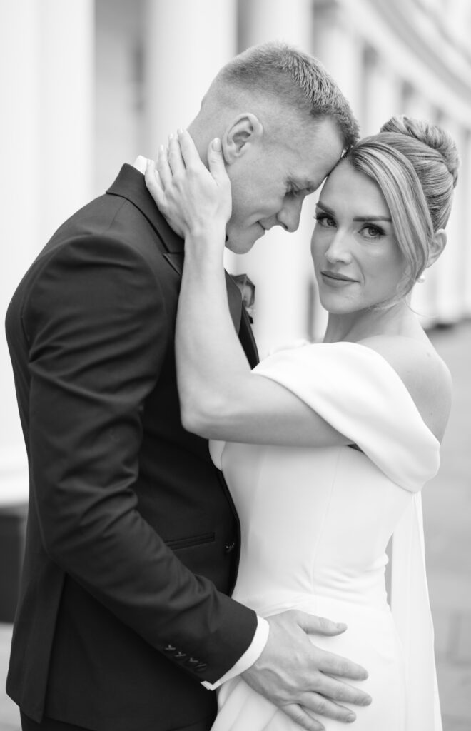 A bride in an off-the-shoulder wedding dress embraces a groom in a suit, gently holding his face as he leans his forehead against her. She looks toward the camera with a soft, serene expression. The image is in black and white.