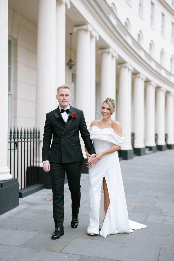 A bride in an off-shoulder white gown with a thigh slit walks hand in hand with a groom in a black tuxedo with a red rose boutonniere. They smile as they stroll outside a grand building with tall white columns.