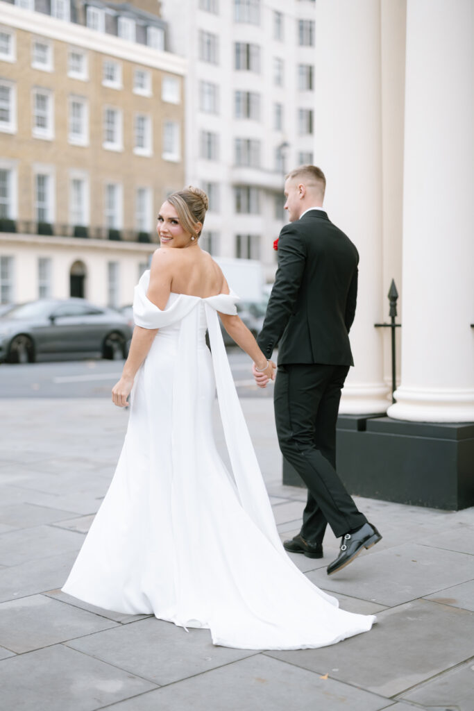 A bride in a white off-the-shoulder gown holds hands with a groom in a black suit as they walk outside on a city sidewalk, with the bride looking back and smiling. Tall buildings and parked cars are in the background.