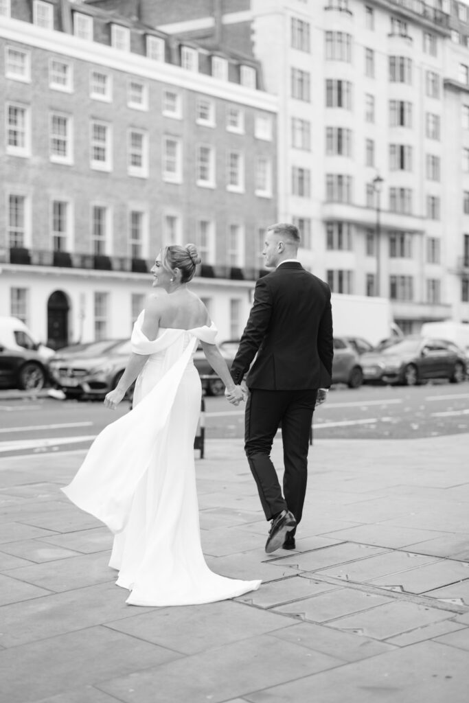 A bride in an off-shoulder white gown and a groom in a black suit walk hand in hand on a city sidewalk, with tall buildings and parked cars in the background.