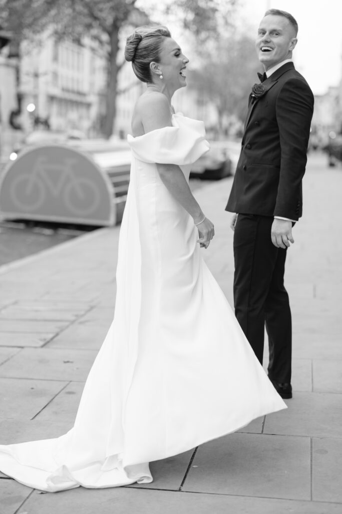A bride in an off-the-shoulder white gown and a groom in a black tuxedo stand outside on a city sidewalk, both smiling and laughing. The background shows blurred buildings and a bike rack.