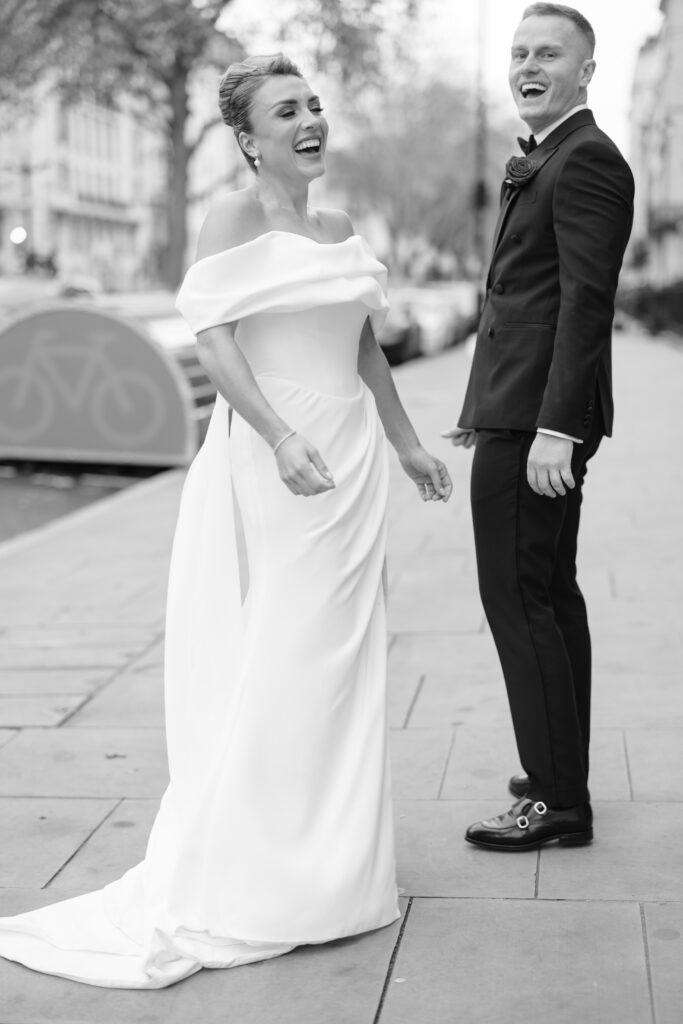 A smiling bride in an off-the-shoulder wedding gown and a groom in a tuxedo laugh together while standing on a city sidewalk, surrounded by parked cars and trees. The photo is in black and white.