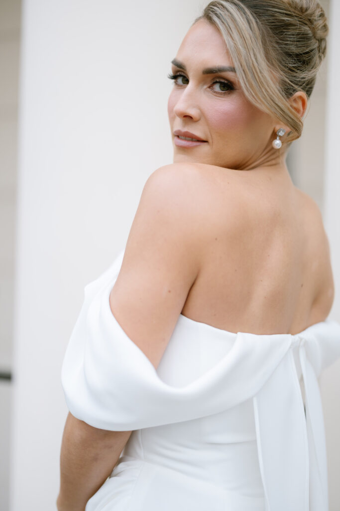 A woman in an off-the-shoulder white dress with an elegant updo looks back over her shoulder, displaying natural makeup and pearl earrings against a soft, neutral background.