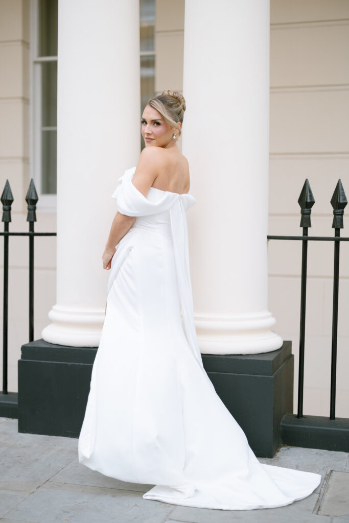 A woman in an off-the-shoulder white gown stands outdoors, looking over her shoulder. She has an elegant updo hairstyle and is posed in front of white columns and a black iron fence.