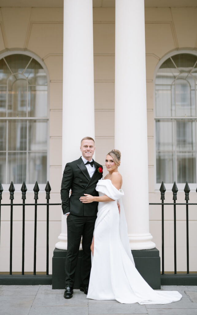 A bride in an off-the-shoulder white gown stands next to a groom in a black tuxedo. They pose together in front of tall white columns and large arched windows, with a black iron fence in the background.