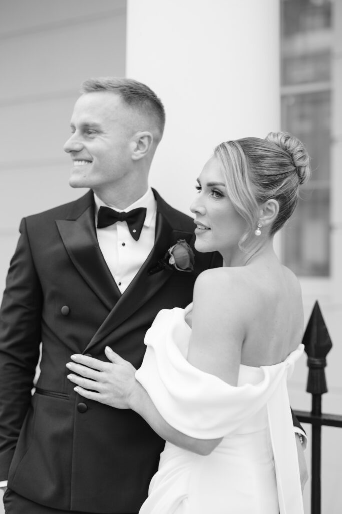 A black and white photo of a bride and groom in formal attire. The groom wears a tuxedo and smiles while looking away; the bride, in an off-shoulder dress, stands beside him with her hand on his chest, also gazing away.