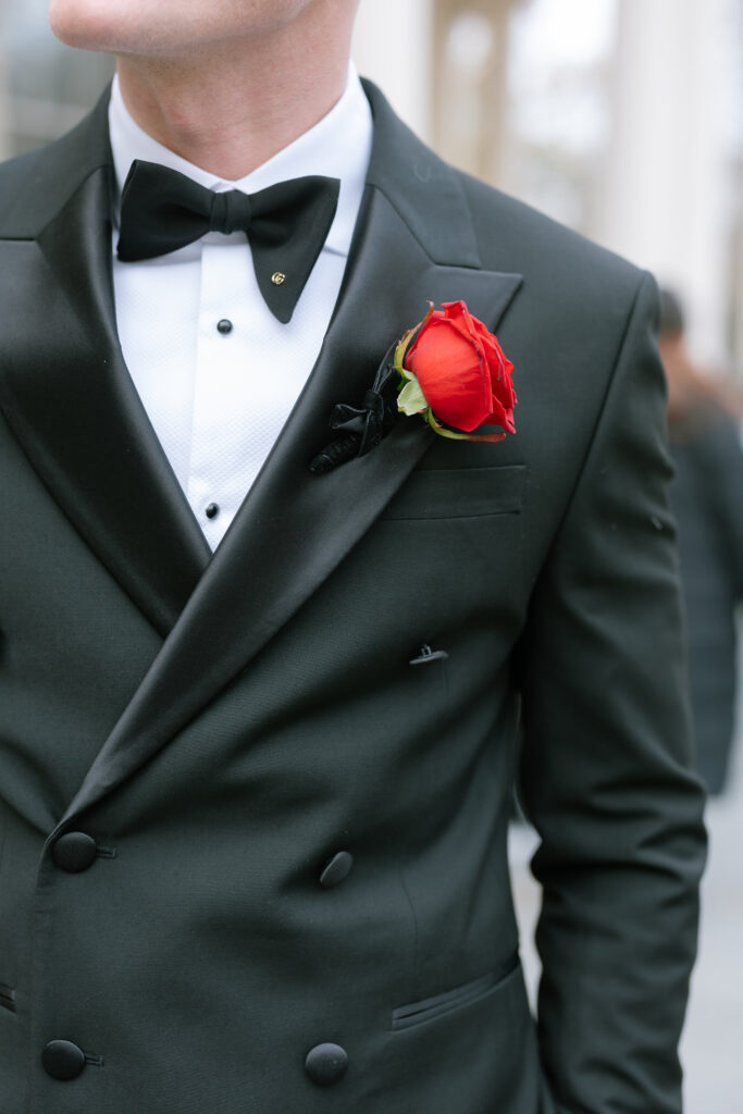 A man wearing a black tuxedo, white dress shirt, and black bow tie, with a red rose boutonniere pinned to his lapel. His face is partially out of frame. The background is blurred.