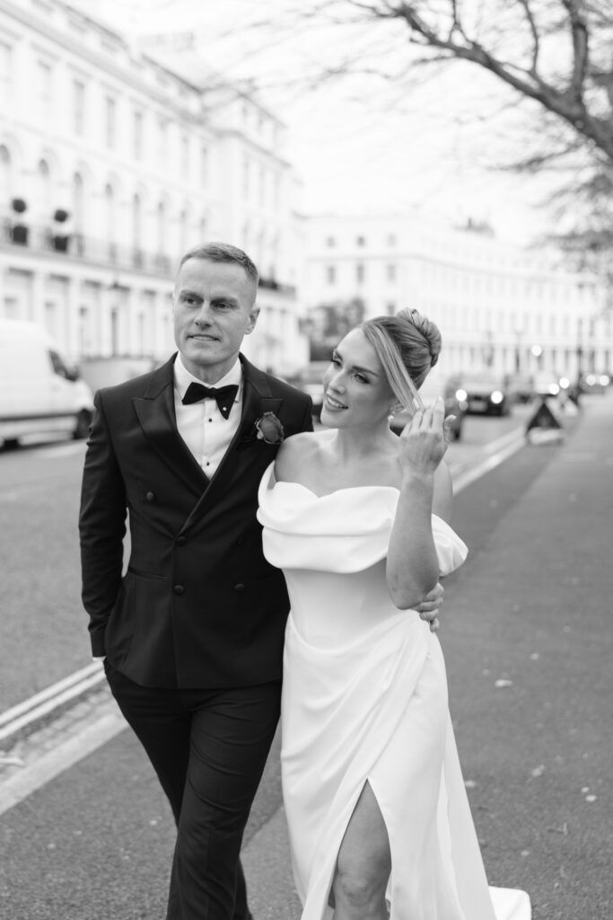A bride and groom walk together on a city street. The groom wears a black tuxedo while the bride wears an off-the-shoulder white dress with a thigh-high slit, smiling and touching her hair. The scene is in black and white.