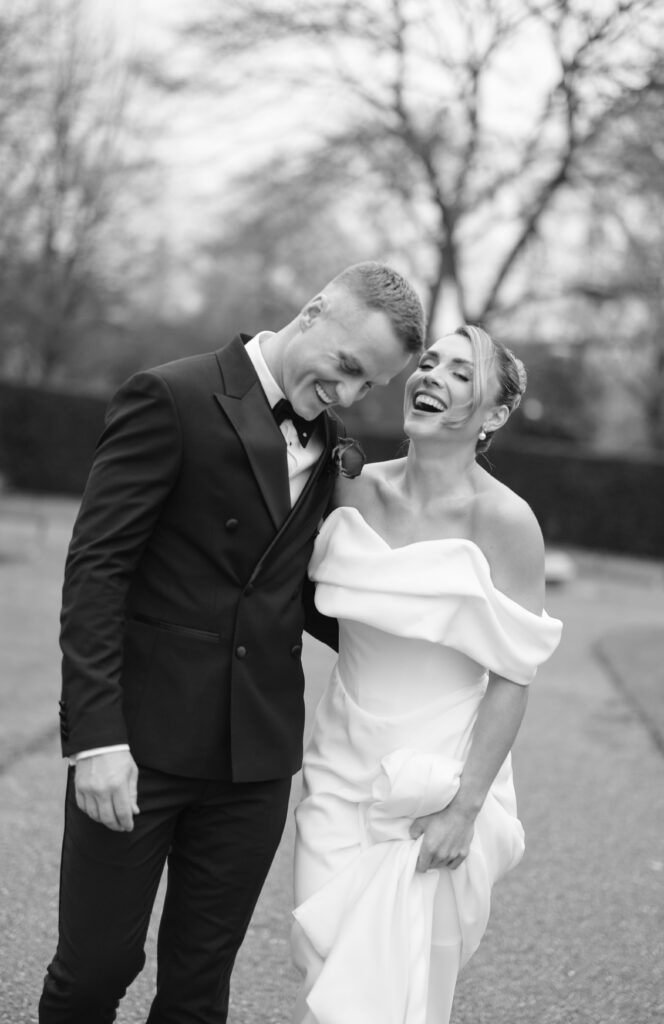 A newlywed couple, dressed in a tuxedo and an off-the-shoulder wedding gown, walk outside together, laughing joyfully on a paved path with trees in the background. The photo is in black and white.