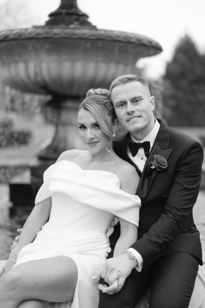A bride in an off-the-shoulder white gown and a groom in a black tuxedo sit closely together, holding hands, in front of a large outdoor fountain. The photo is black and white.