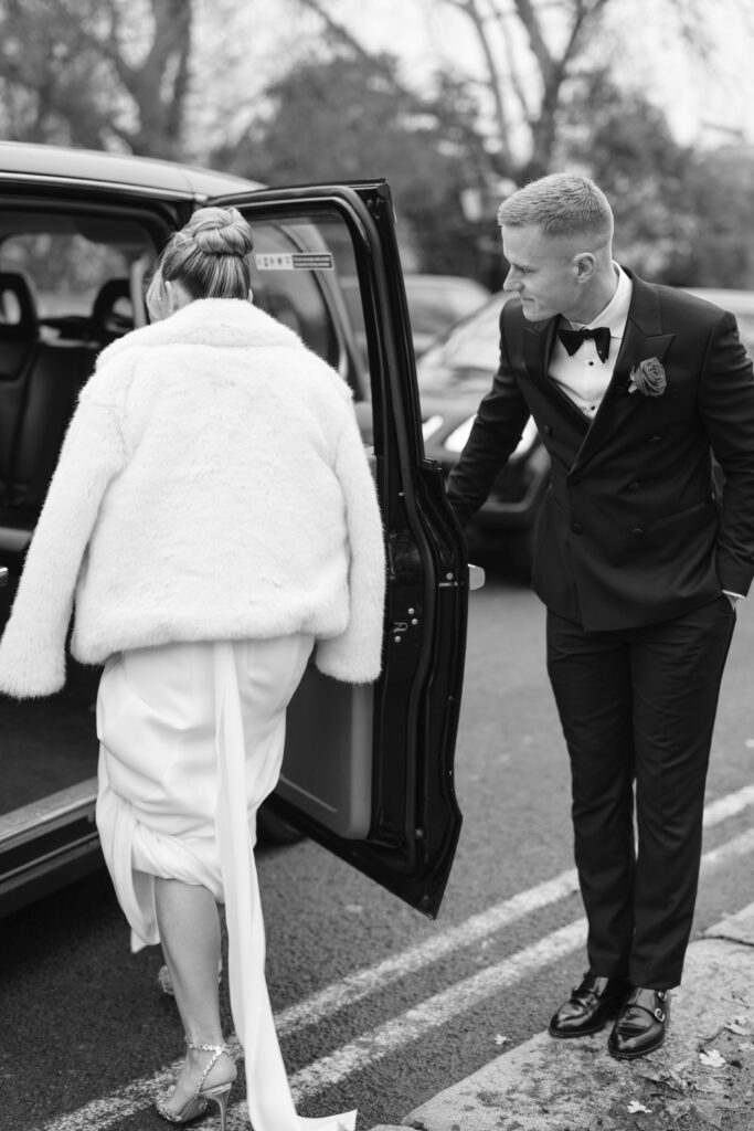 A bride in a white dress and fur jacket steps into a car as a groom in a black tuxedo stands nearby, holding the door open for her. The scene takes place on a street, captured in black and white.