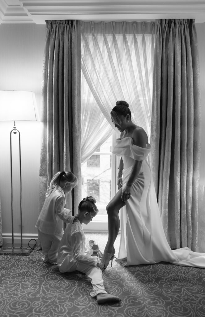 A bride in an off-shoulder gown stands by a window as two children in pajamas help her with her high-heeled shoe in a softly lit room with long curtains.