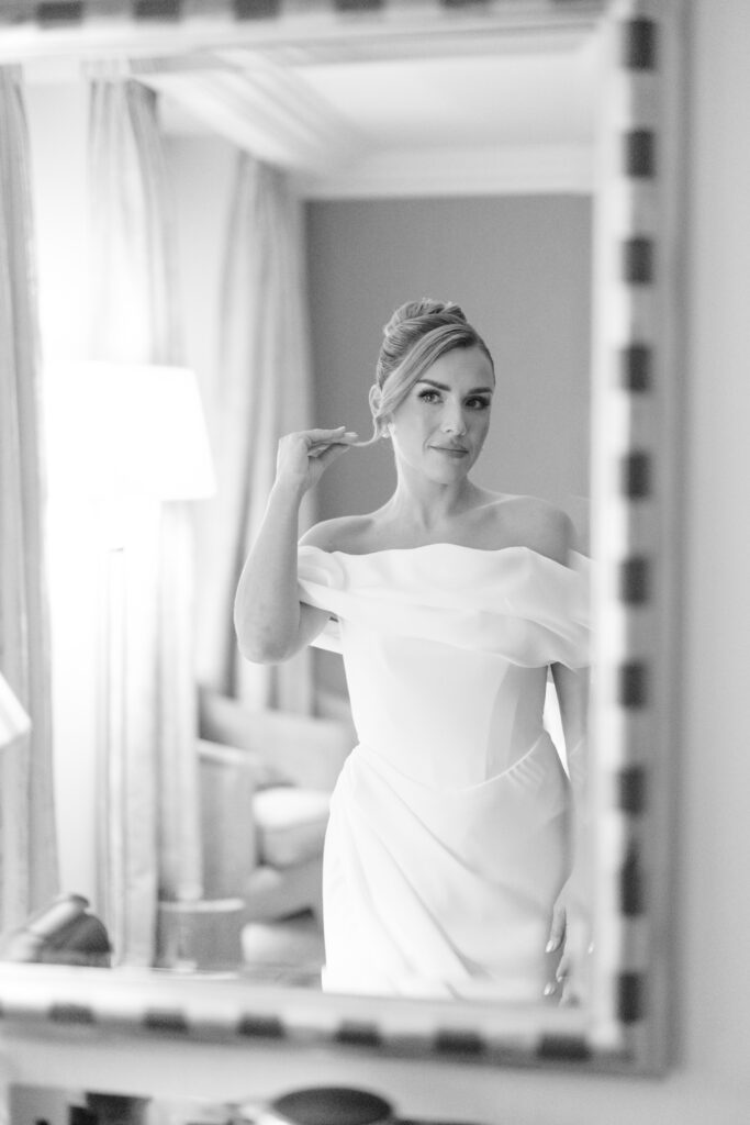 A woman in an off-the-shoulder white dress stands in front of a mirror, adjusting her earring. The scene is elegant and softly lit, with curtains and a lamp visible in the background.