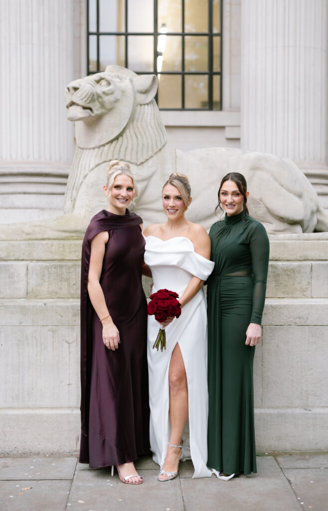 Three women stand in front of a stone lion statue and tall columns. The center woman wears a white off-the-shoulder gown and holds red roses, flanked by women in dark purple and green dresses. All are smiling at the camera.
