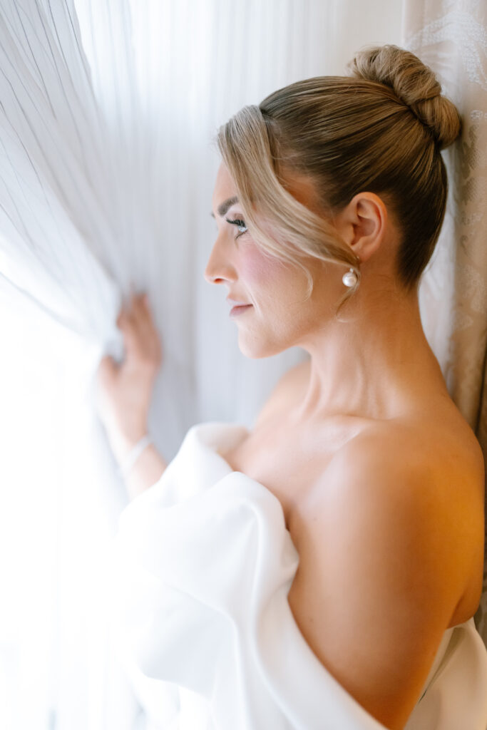 A woman in an elegant off-the-shoulder white dress stands by a window, gazing outside. Her hair is styled in a neat bun, and she wears pearl earrings. Soft natural light illuminates her profile.