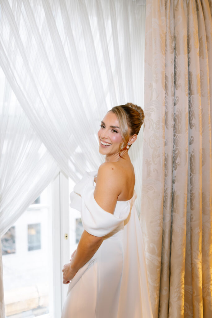 A smiling woman in an off-the-shoulder white dress stands by a window with sheer curtains, looking over her shoulder. Elegant drapes and soft natural light create a warm, cheerful atmosphere.